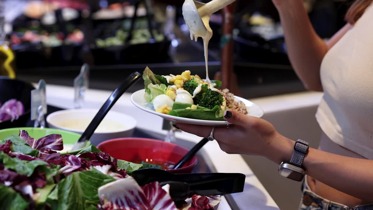 Woman adds creamy dressing to mixed greens and vegetables at self-serve salad bar, indoor lighting