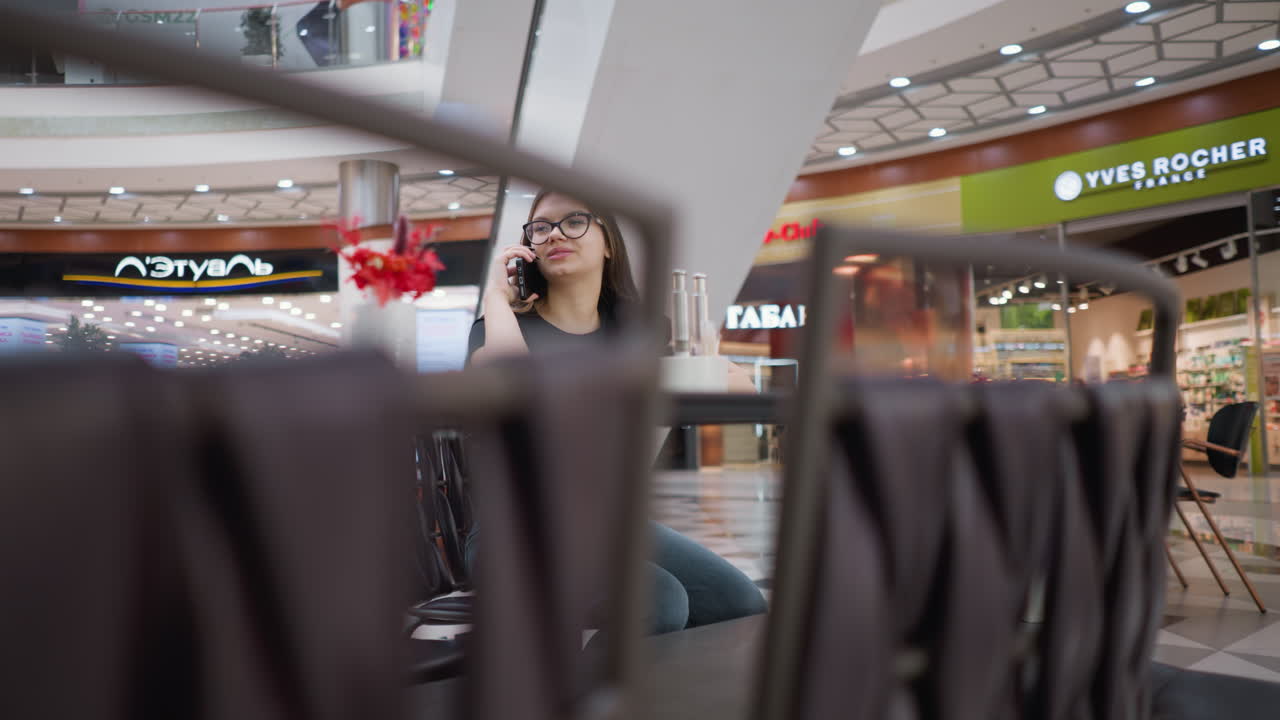 아름다운 구조의 쇼핑몰에서 전화 통화를 하는 안경을 입은 여인과 함께 배열 된 의자의 클로즈업, 도시 소매 분위기를 보여줍니다.