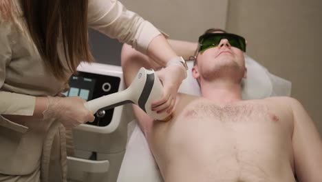 a professional laser technician performs hair removal treatment on a male client's underarm area in a clinical setting, with both wearing safety gear