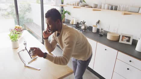 happy african american man leaning on countertop in kitchen, using tablet and talking on smartphone