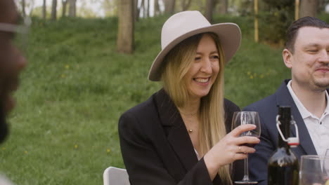 mujer rubia sosteniendo una copa de vino tinto y riendo mientras se sienta a la mesa con amigos durante una fiesta al aire libre en el parque