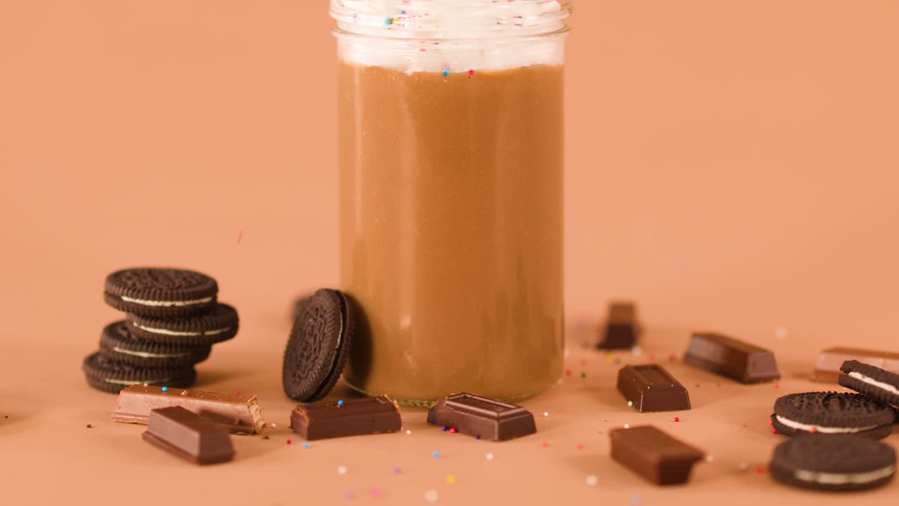 Chocolate milkshake topped with whipped cream, surrounded by cookies, chocolate, and colorful sprinkles