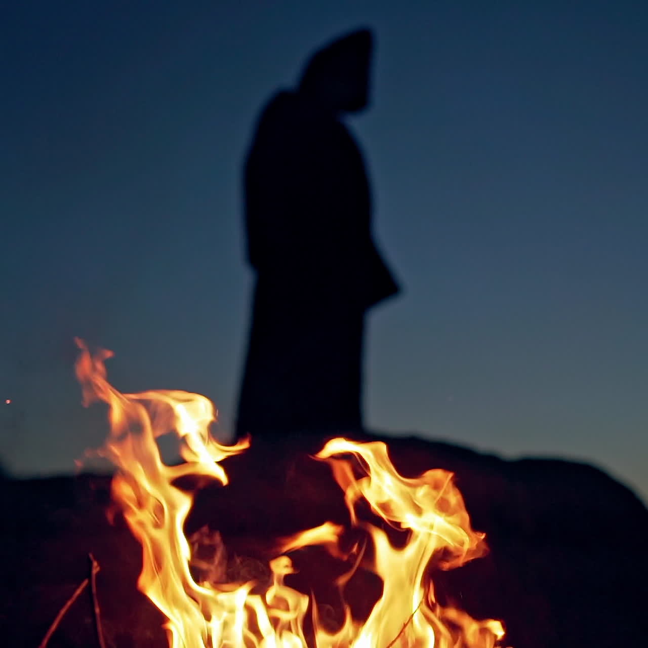 Fire is burning on the dark background with black figure. Scary atmosphere on Halloween. Ghost in black behind the vivid flame outdoors.