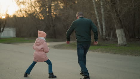 a man in a green jacket and a little girl in a pink jacket, both wearing blue jeans, are seen joyfully jumping and playing together outdoors. the scene captures a playful moment of bonding and happiness