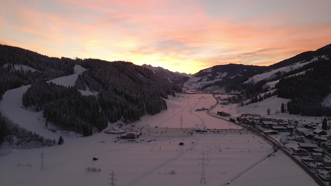 toma aérea de un pueblo de montaña cubierto de nieve, camino sinuoso a través del valle durante la puesta de sol