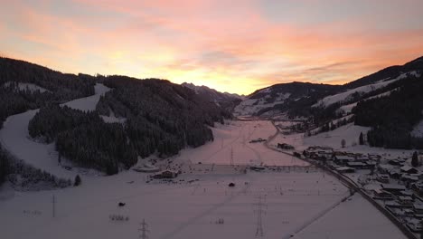 Aerial-shot-of-a-snow-covered-mountain-town,-winding-road-through-the-valley-during-sunset