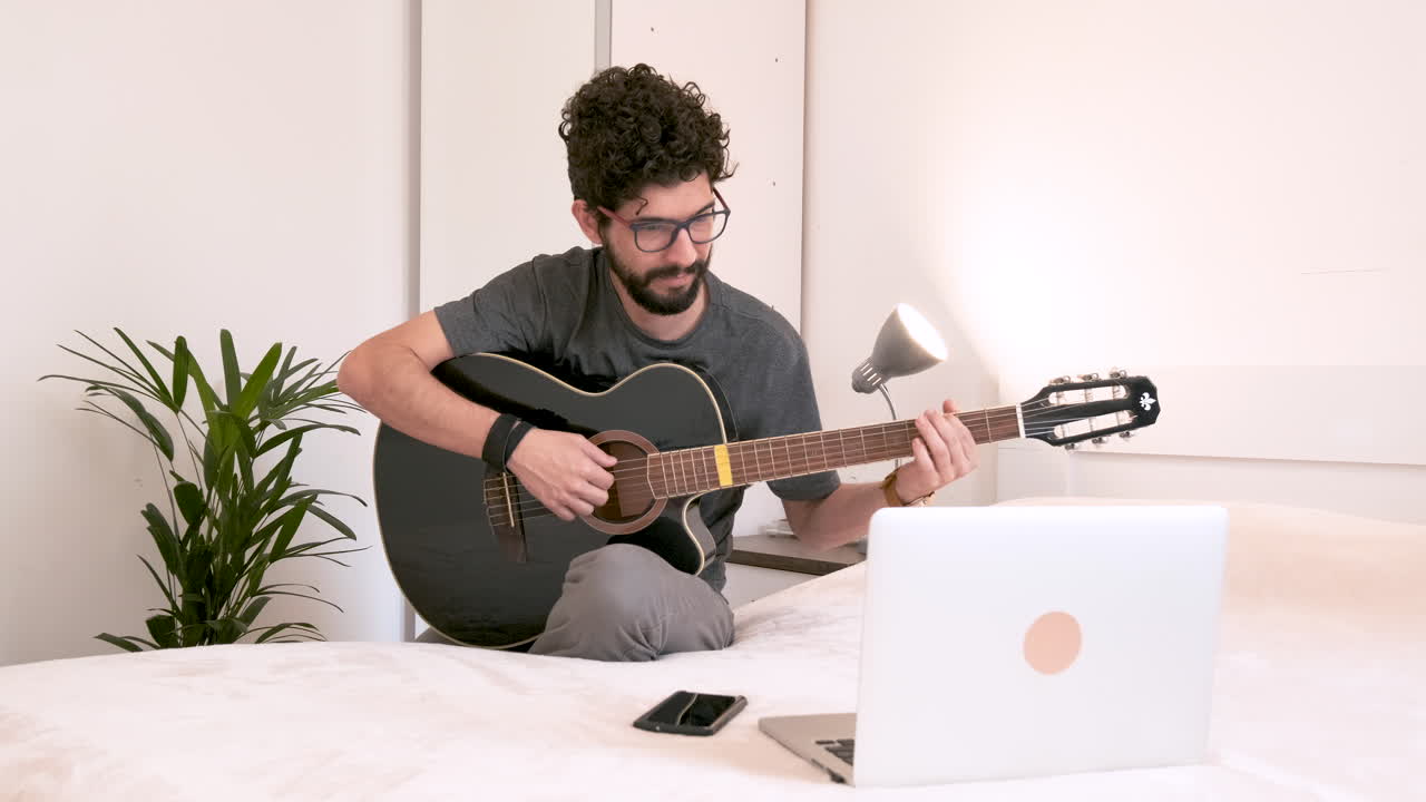 hombre tocando la guitarra en la habitación frente a la laptop en un fondo claro, aislado