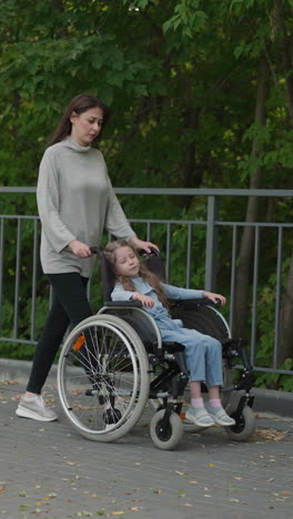 little girl rests in wheelchair pushed by mother in city park with lush green trees. woman takes daughter with cerebral palsy for spring walk