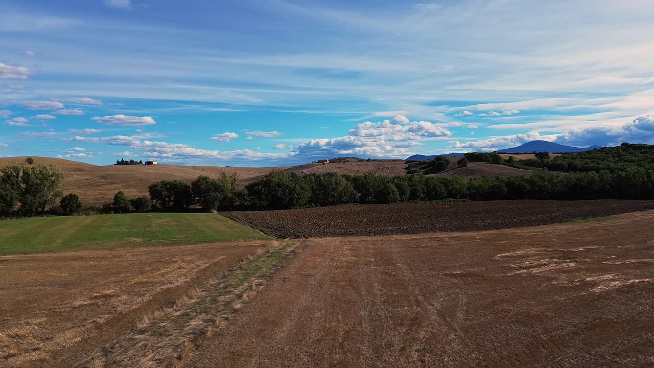 Breathtaking view of the Tuscan countryside showcasing rolling hills, farmland, trees, and a vibrant blue sky with scattered clouds.