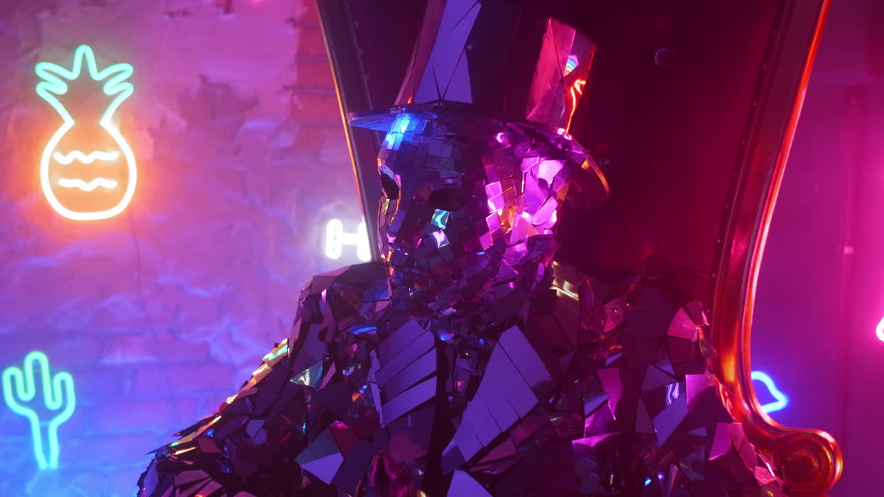 un hombre con un traje de metal plateado de acero se sienta en una silla y sacude la cabeza a la música en la luz de neón.