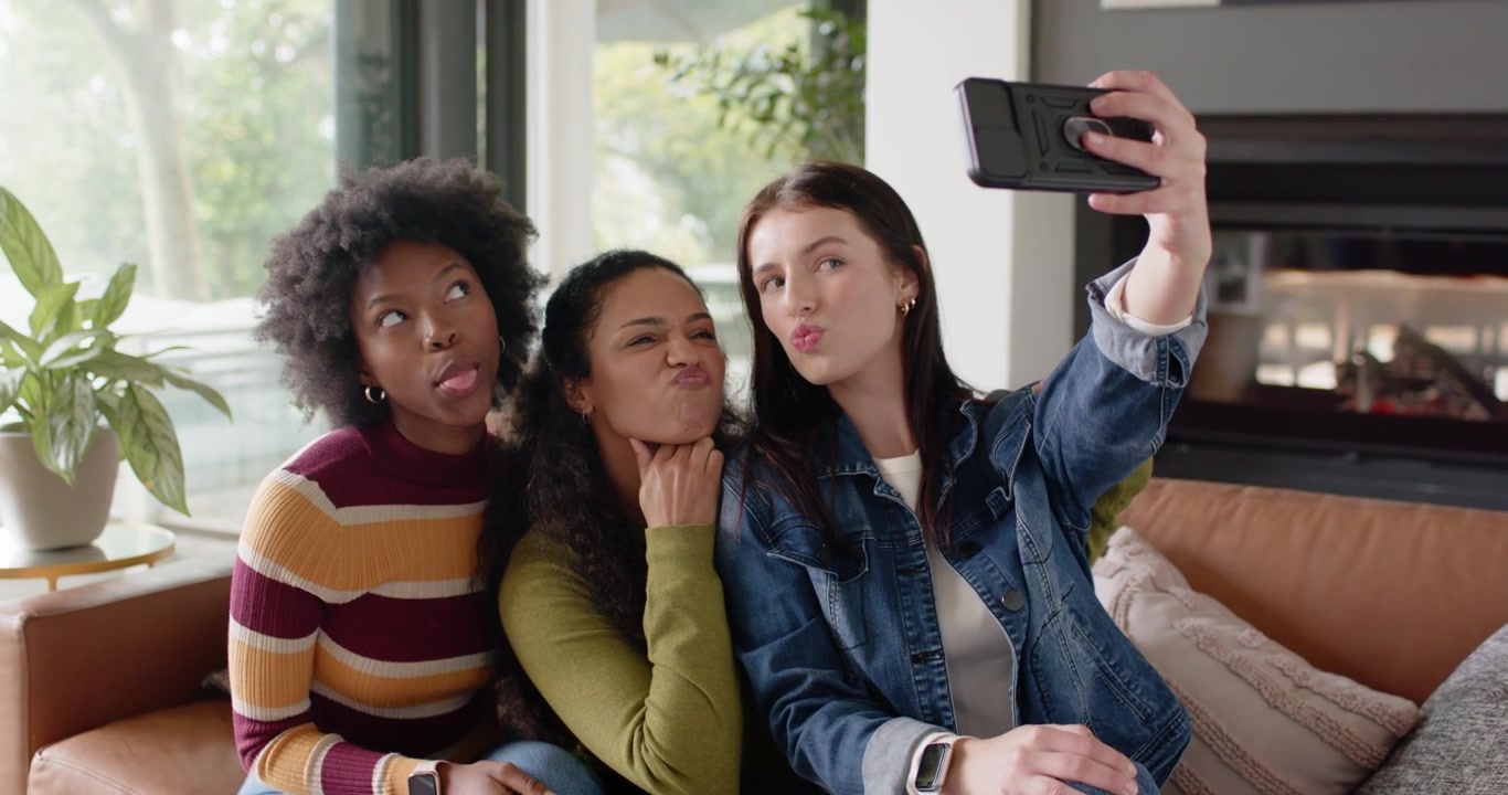 Diverse female friends passing smartphone, reacting and posing for selfie on brown leather sofa