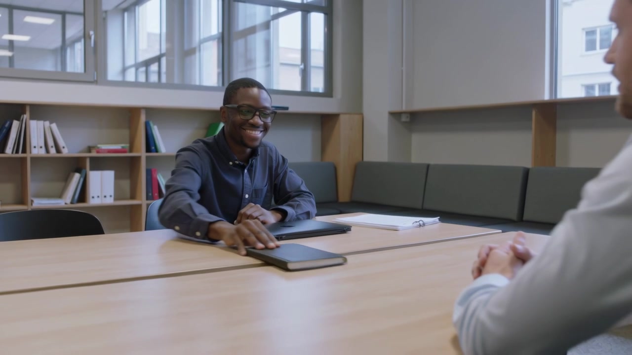 Closing laptop, man with glasses and dark shirt sliding notebook to peer entering room for review