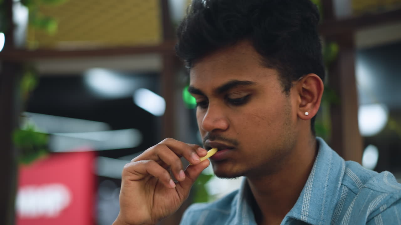 Indian young man taking bite from fries, close up of mouth and hand holding crispy pieces, soft light, subtle expression of enjoyment and focus, background blurred to emphasize texture and action