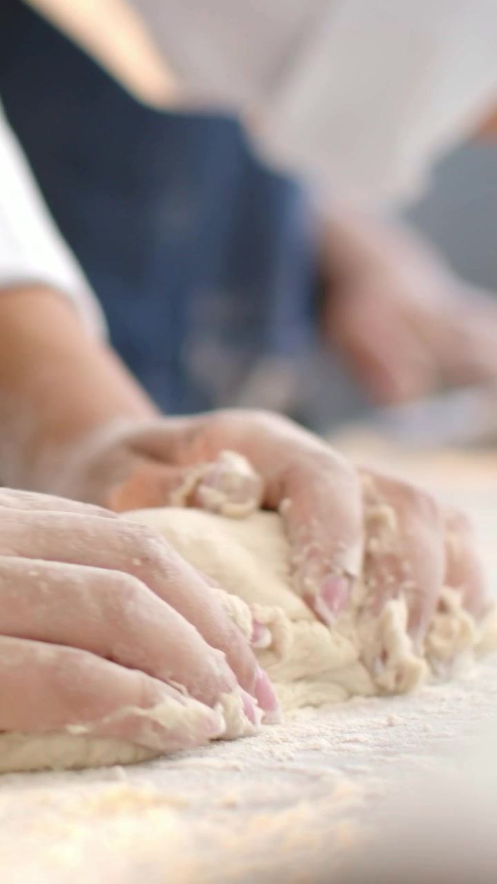 Video verticale: Mani di chef che premono, piegano e modellano una palla di impasto su un piano di lavoro infarinato, con polsino bianco.