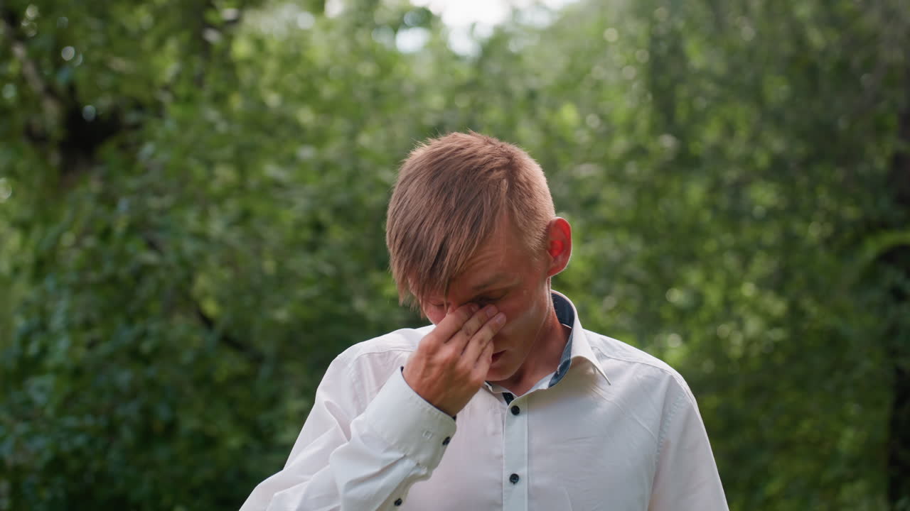 Young man wearing white shirt appears exhausted while adjusting glasses in forest, standing under daylight surrounded by dense greenery, expressing fatigue and tired mood in natural outdoor