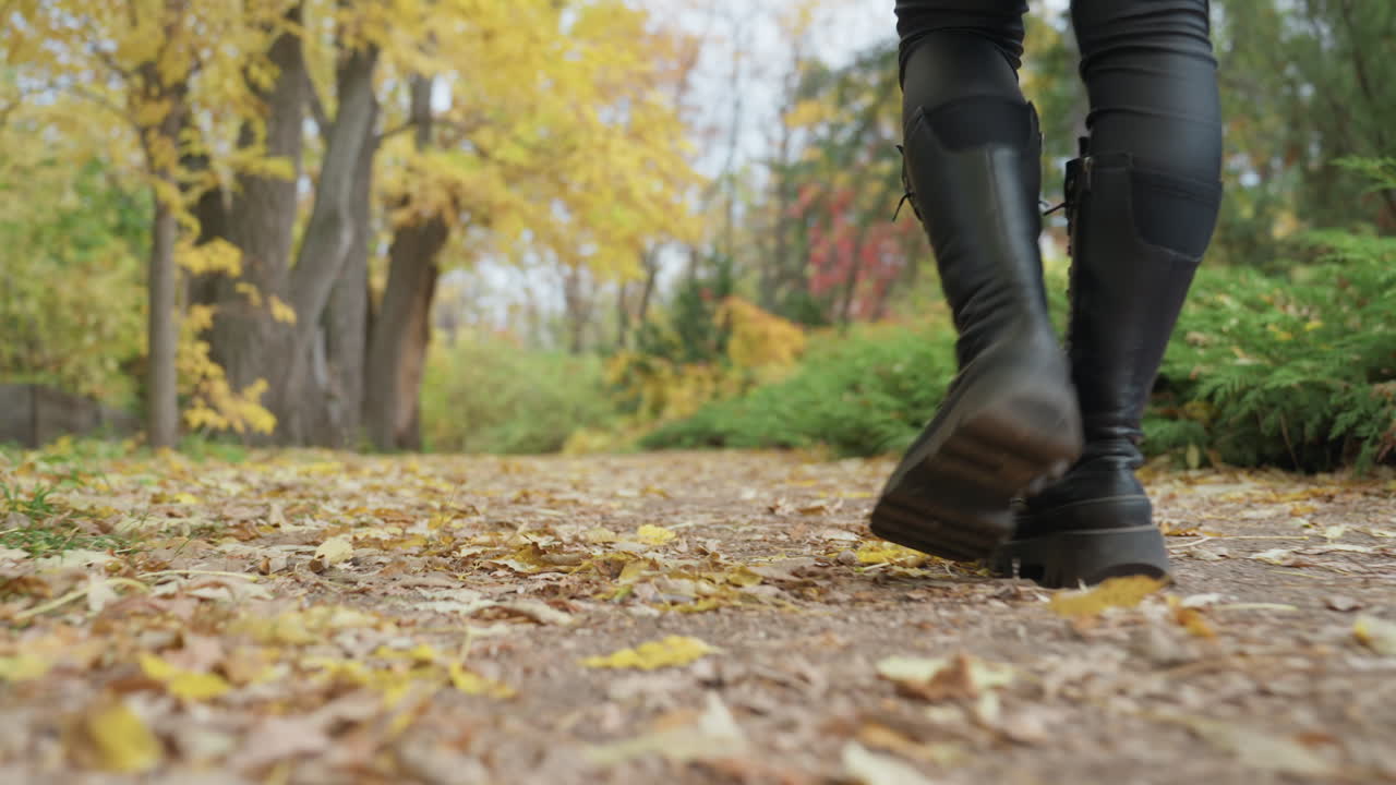 Botas de cuero caminando sobre sendero de hojas, parque otoñal bordeado de follaje amarillo, primeros planos a nivel del suelo de una mujer con pantalones de cuero negros, zancada segura sobre suelo crujiente, ambiente cinematográfico melancólico, indie