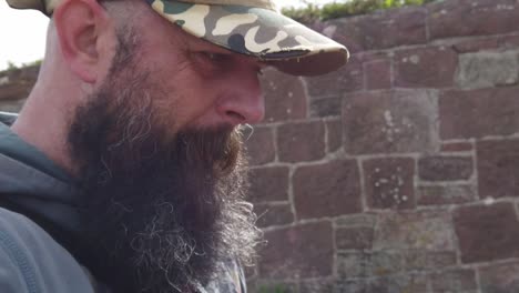 hombre barbudo con gorra de béisbol con expresión preocupada caminando junto a la pared de piedra