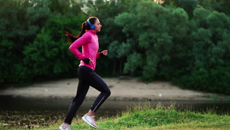 una chica con una chaqueta rosa y pantalones negros corre cerca del río con auriculares preparándose para el maratón