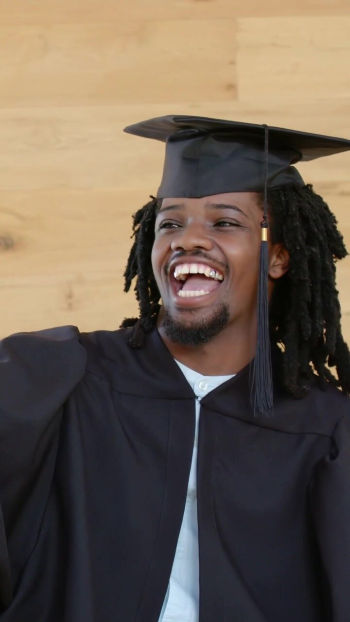 Vertical video: Extending arm, grad in gown taking phone selfie at wood wall, marking grad, tassel