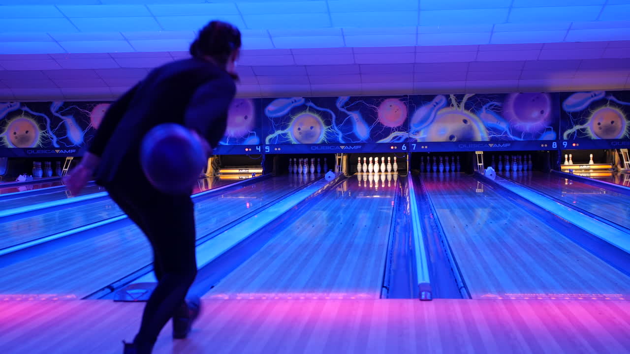 A female bowler throwing a ball down the lane in a neon-lit bowling alley, with brightly colored pins lined up in the distance.