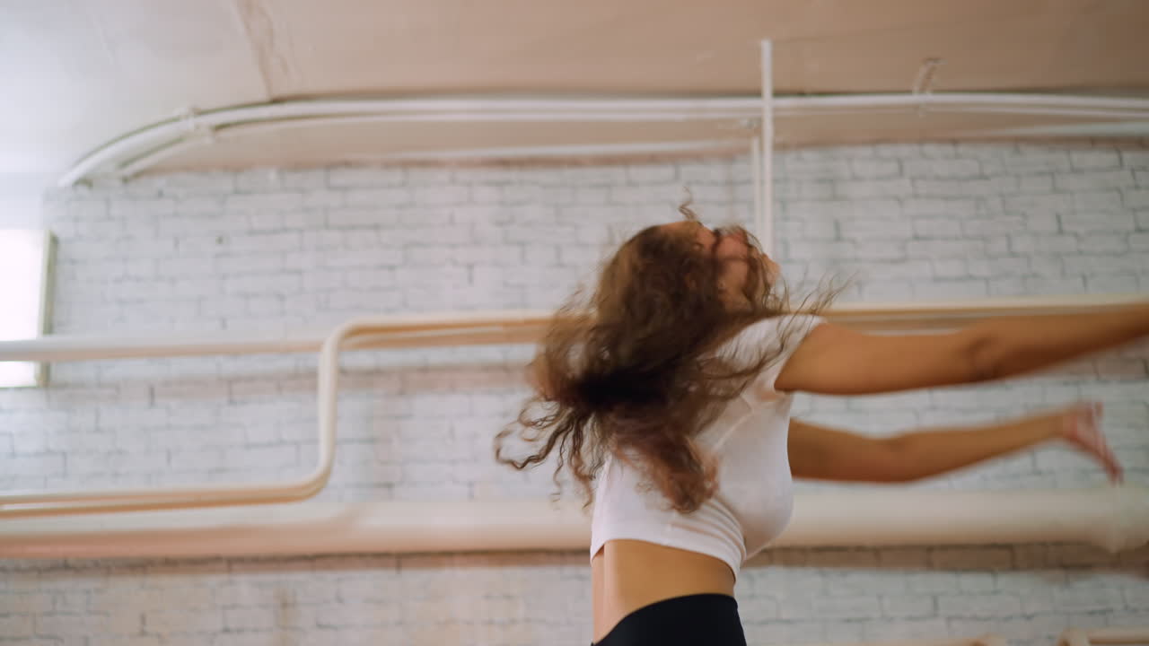 Side view of young woman in white top and black leggings dancing in studio placing hand on waist pushing out chest expressive confident movement athletic rhythm graceful posture energy