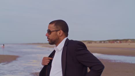 A-male-man-black-latino-model-in-a-suit-walks-on-the-beach-and-takes-off-his-jacket-at-the-seashore-with-sunglasses-in-the-Netherlands,-the-Hague