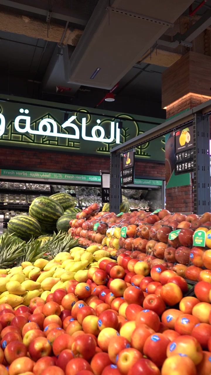Exposição colorida de várias frutas frescas e maduras nas prateleiras da seção de hortifrúti de um supermercado, com maçãs, peras, melancias e abacaxis prontos para compra.