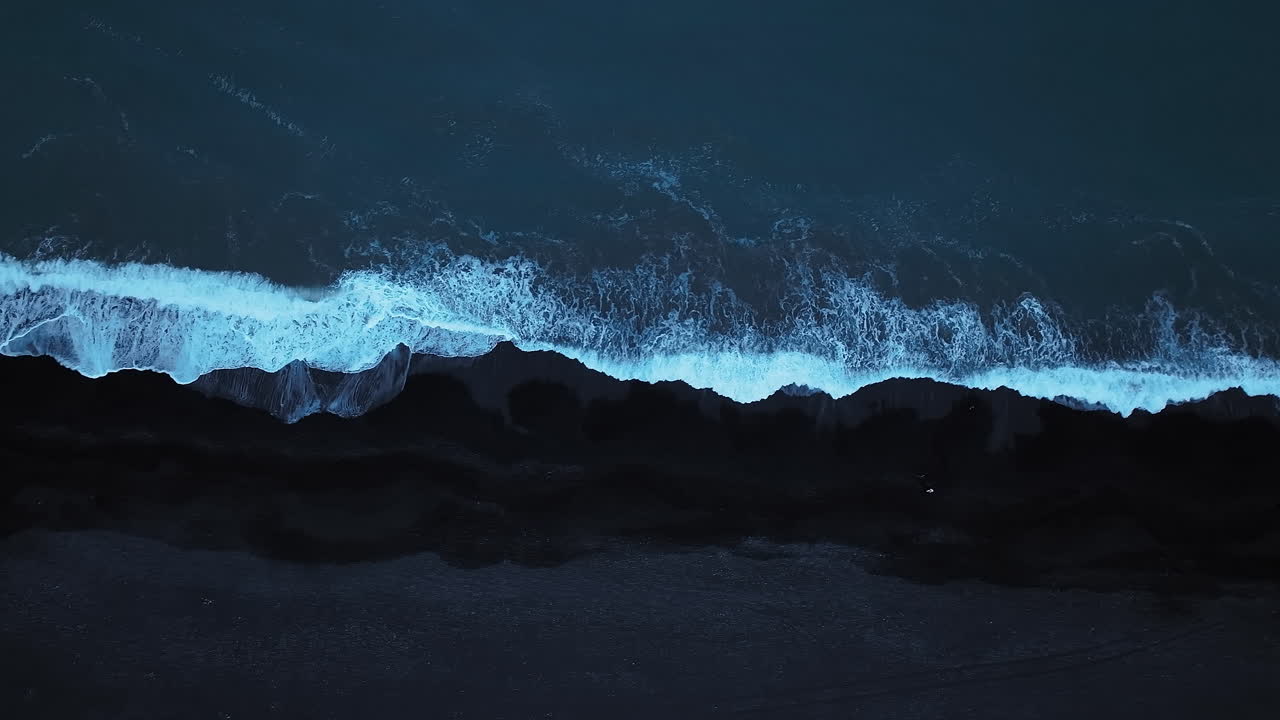 Cold ocean waves lapping a black volcanic sand beach, aerial directly above shot