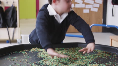 Primary-schoolboy-with-Down-Syndrome-stands-using-a-sensory-play-tub-in-a-classroom,-close-up