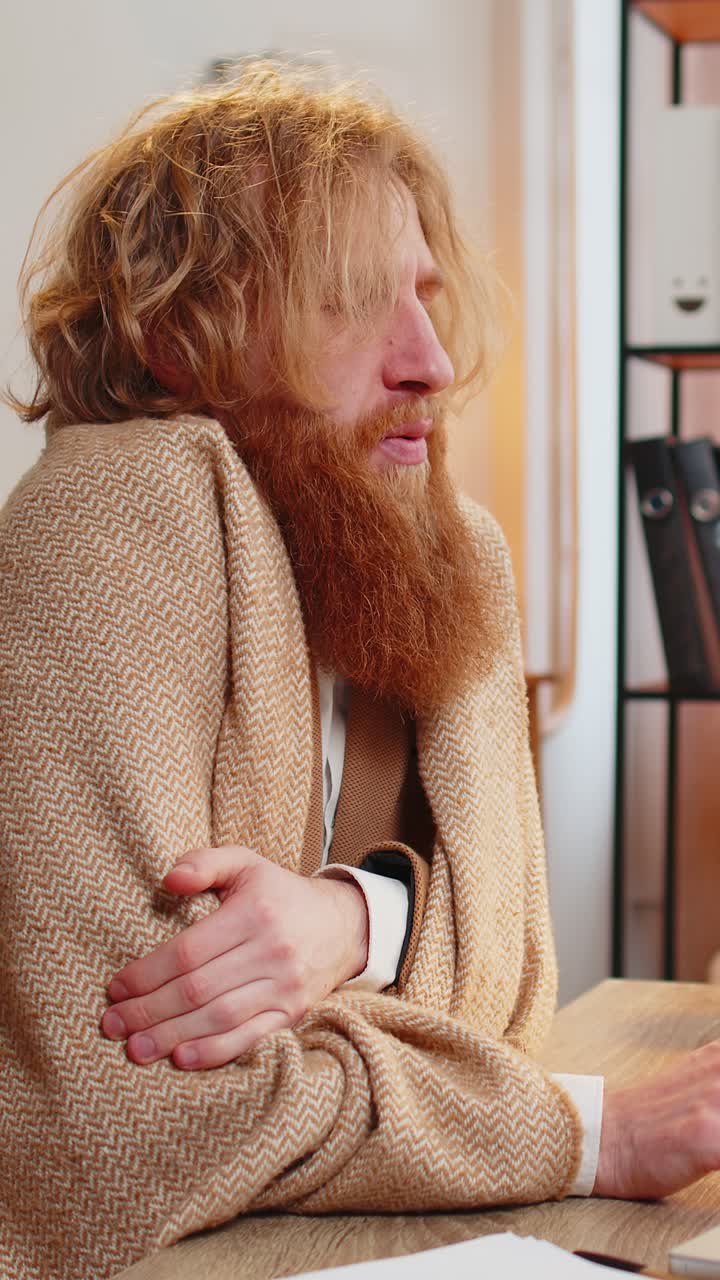 아픈 사업가가가 차가움으로 떨고, 담요로 감싸서 재채기를 하고, 사무실에서 노트북으로 일하고 있다.