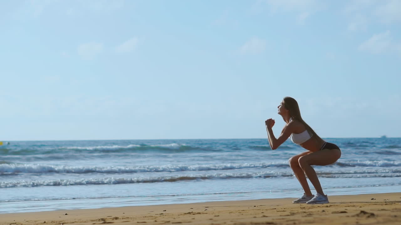 Giovane donna in forma che allena addominali e glutei con un allenamento a corpo libero, eseguendo esercizi di squat in spiaggia. Ragazza sportiva che fa squat come parte di una vita attiva e in forma. Ripresa con steadicam.