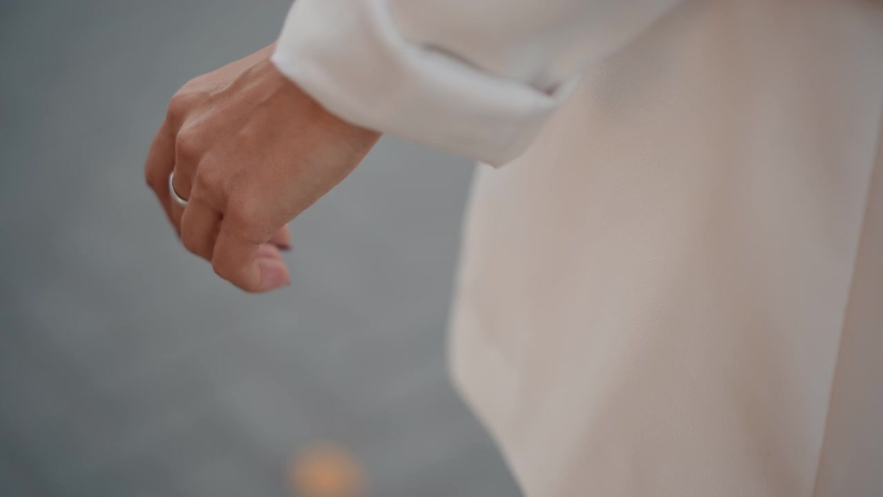 Beskåret bevægelsesskud, der viser underarm og afslappet hånd ved siden af en lys langærmet frakke eller kjole med oprullet manchet, stofkant og ærme, der svajer blidt, som om man går, eller en brise bevæger tøjet; lav