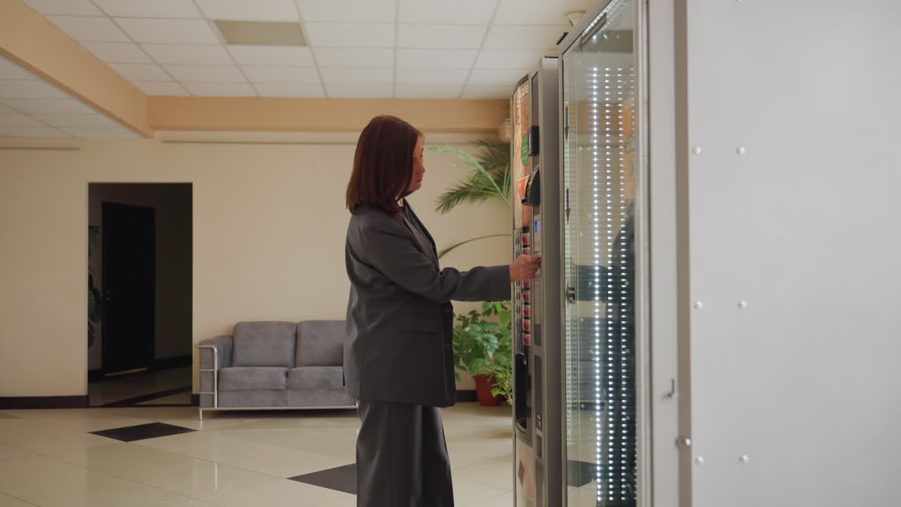 Professional businesswoman selecting snack from vending machine in office lobby, wearing suit, taking a break during workday in modern office building, showing casual moment in corporate setting