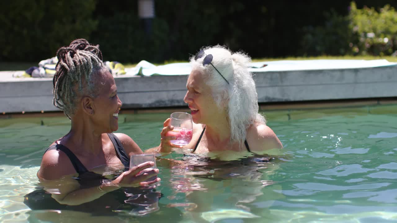 Due donne in costumi da bagno interi che brindano con bevande rosa senza stelo a bordo piscina di casa, socializzando, con occhiali da sole.