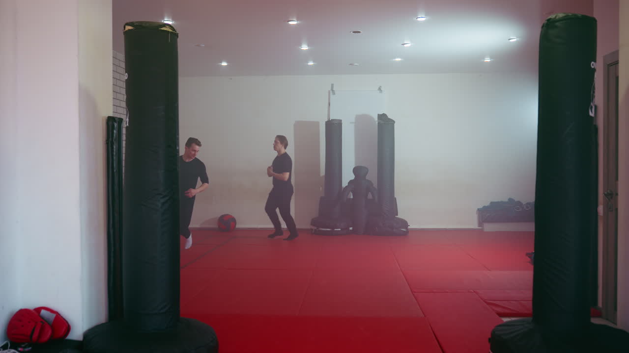 Two fighters in black sportswear jogging on red training mat inside martial arts gym, preparing for combat practice near punching bags, focusing on movement, stance, and readiness for sparring session