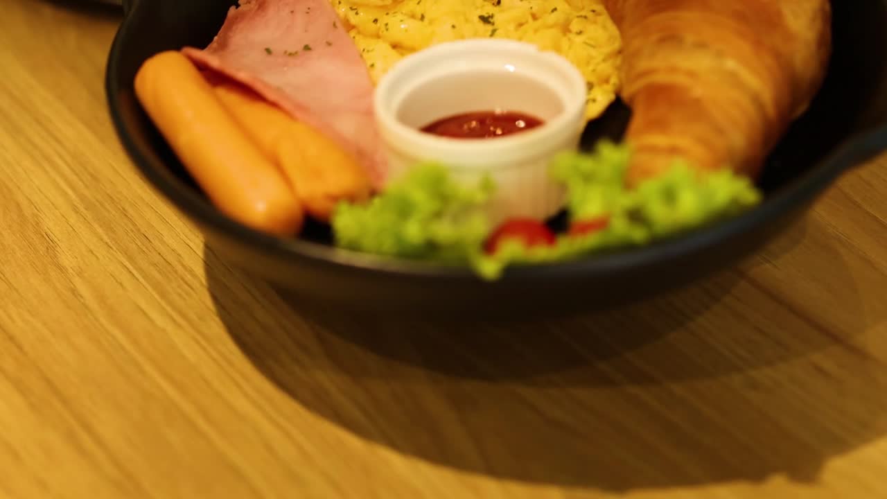 A close-up of a breakfast plate featuring scrambled eggs, croissant, ham, sausages, and salad on a wooden table.