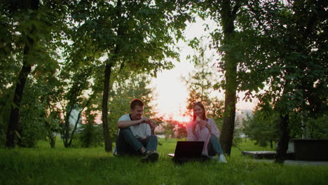 two students sitting on grass outdoors with laptop in front, making a toast with bread in hand, as sunlight filters through trees in the background