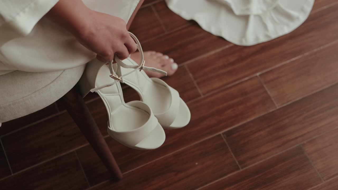 Close up of bride’s hand holding elegant white high heel shoes on wooden floor indoors