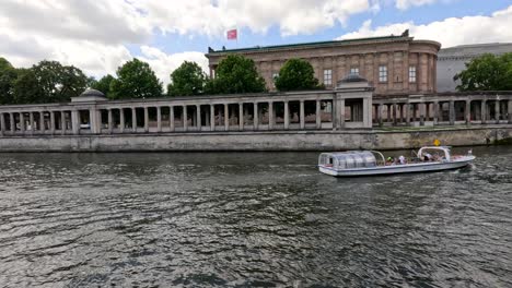 a sightseeing boat glides along the spree river in berlin, passing a neoclassical museum under daylight with steady camera framing and soft natural lighting