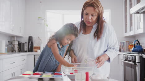 young girl making cakes with her grandmother, filling cake forms with cake mis using spoon, close up