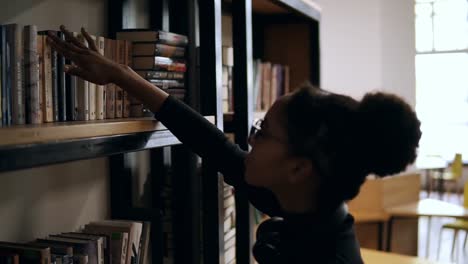 close up side view of the african american young pretty curly woman looking for the books on the shelves of library or a book store. bright window light on the background