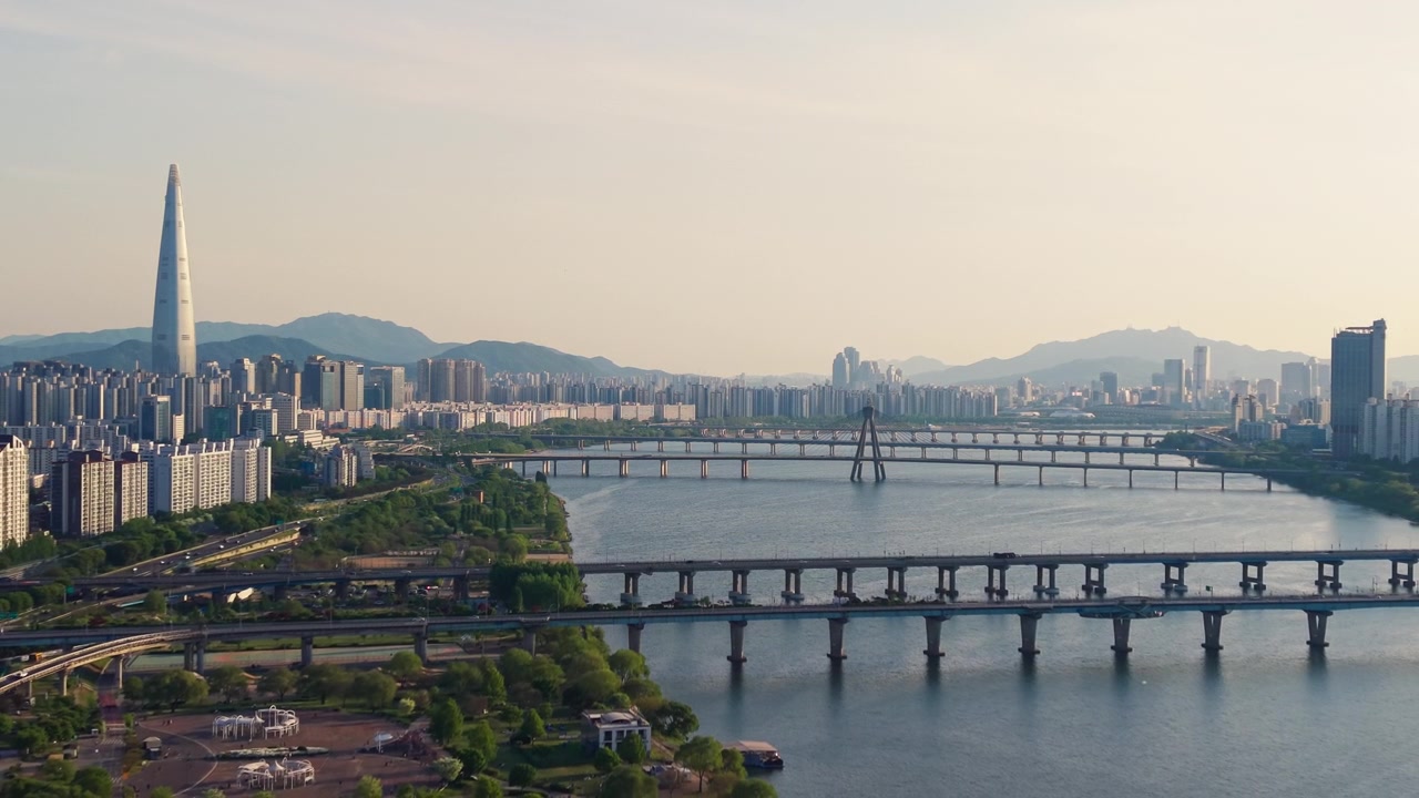 Una panoramica aerea all'indietro sul Parco Gwangnaru Hangang rivela i ponti Gwangjin e Cheonho che attraversano il fiume Han, con la Lotte World Tower e lo skyline dorato e velato di Seul al tramonto.