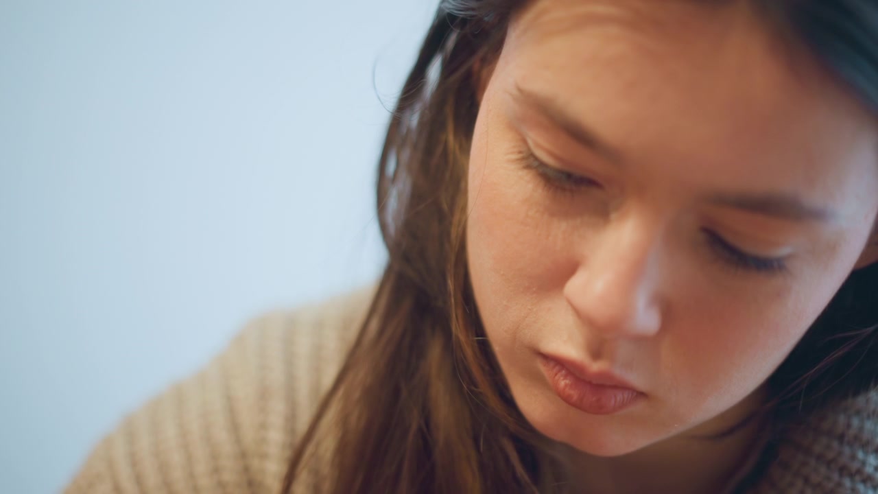 Close up of woman with long brown hair wearing beige knitted sweater softly lit against light blue background. Hair texture and knit pattern details are visible. Camera remains steady