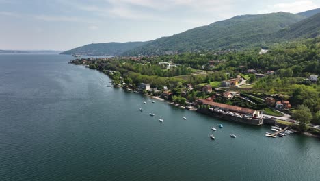 Aerial-slow-orbit-shot-over-Lago-Maggiore-green-hill-shoreline-with-tile-roofed-buildings,-boats-at-anchor-on-a-beautiful-sunny-day-and-clear-blue-sky