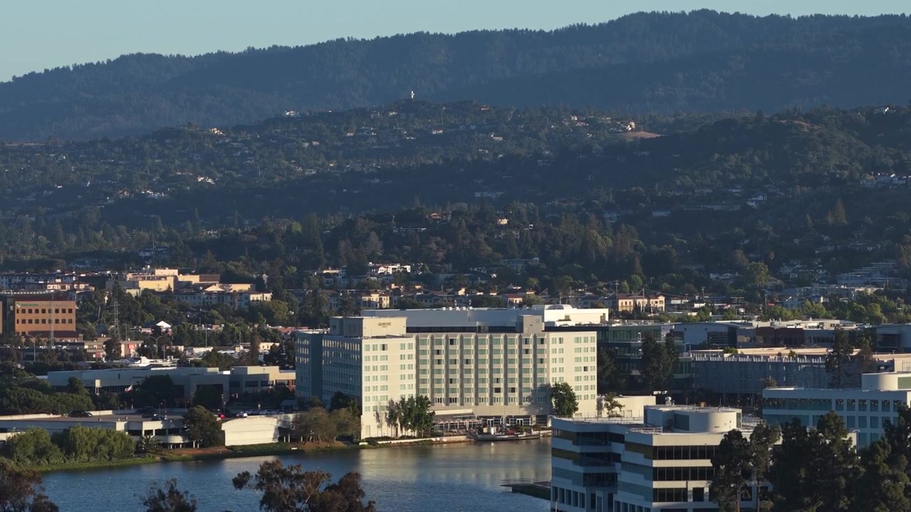 Vista aerea con drone in ascesa che mostra il quartiere di Redwood Shores a Redwood City, California, con i suoi caratteristici edifici per uffici, un lago tranquillo e montagne panoramiche visibili in lontananza.