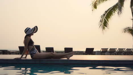 Slow-motion-on-the-silhouette-of-Asian-model-woman-sitting-at-the-edge-of-the-swimming-pool-on-sunset-in-Thailand-resort,-side-view