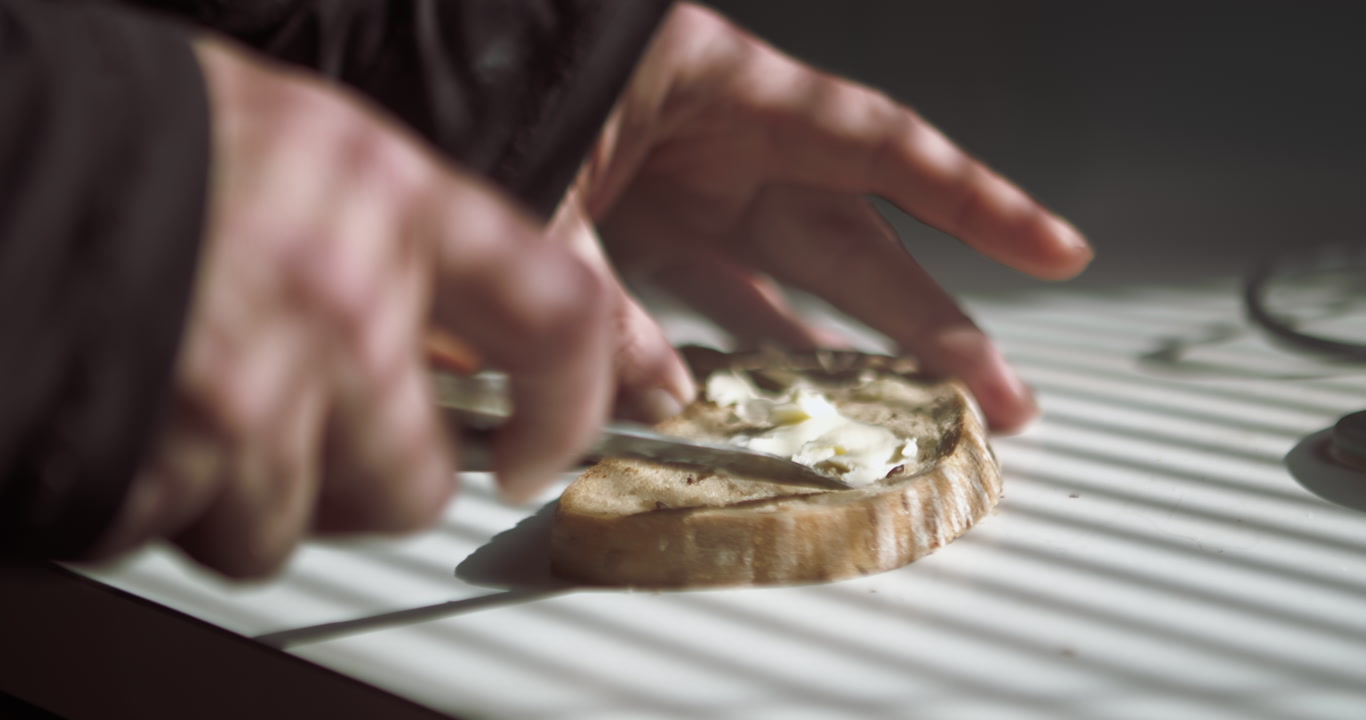 Detailed close-up of hands spreading butter on toast in morning sunblind shadows. Ideal for breakfast commercials, healthy lifestyle vlogs, and domestic routine sequences.