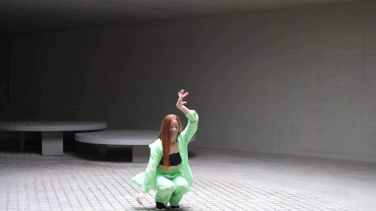 A young woman dressed in a green suit is energetically dancing in a modern concrete setting