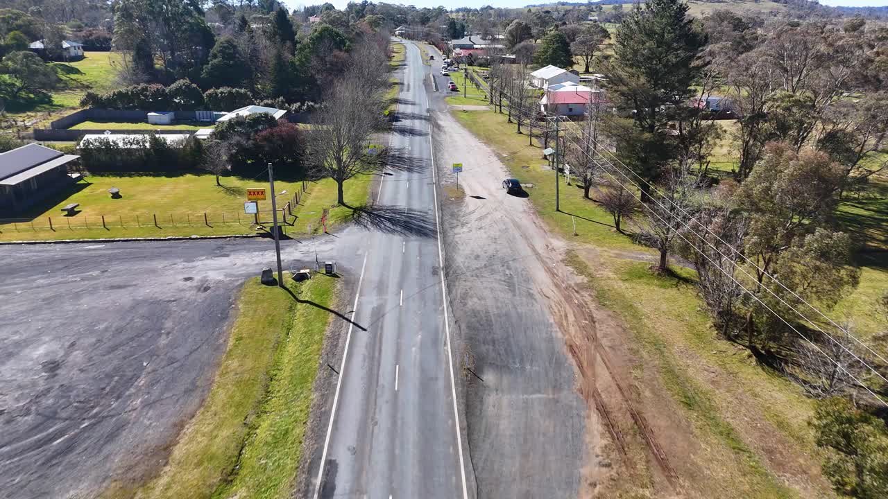 Drone footage smoothly tracks above a quiet rural road, crossing a bridge over a stream and passing houses, parking lots, and green spaces in bright daylight