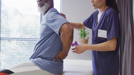 diverse female physiotherapist and senior male patient in hospital physical therapy session on back